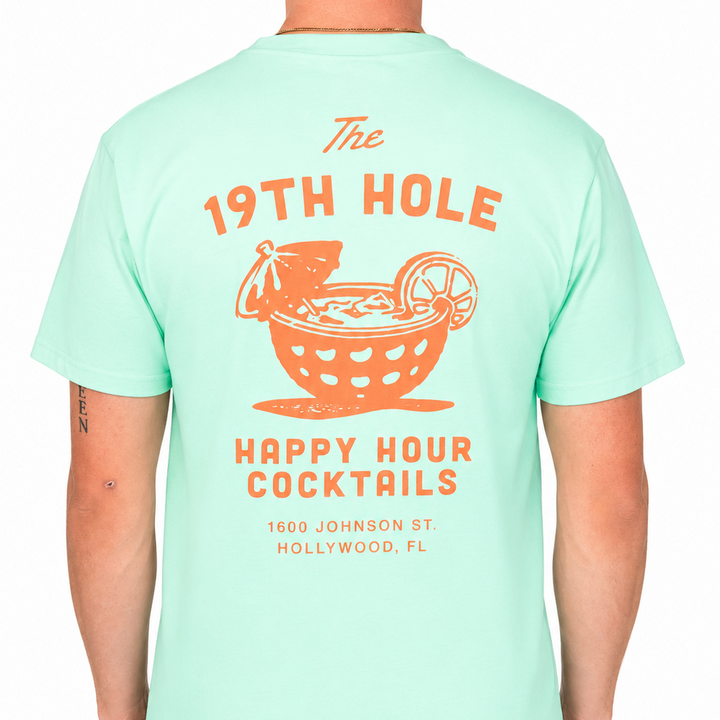 Back view of a male model wearing a mint green graphic t-shirt featuring a large coral “The 19th Hole Happy Hour Cocktails” design with a cocktail-in-a-golf-ball illustration and Hollywood, FL text. Studio product image on a clean white background.