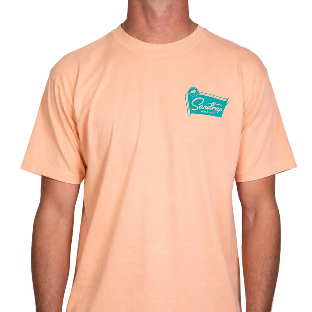 Male model wearing a peach Sandtrap T-shirt featuring a small teal Sandtrap Après Golf logo on the left chest on plain white background.