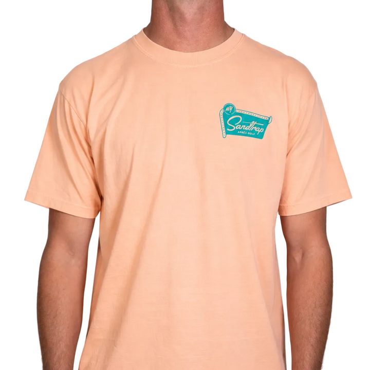Male model wearing a peach Sandtrap T-shirt featuring a small teal Sandtrap Après Golf logo on the left chest on plain white background.