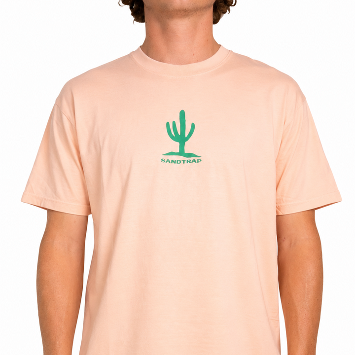 Male model wearing a peach Sandtrap T-shirt featuring a small green cactus logo centered on the chest. The relaxed-fit tee is shown against a clean studio background, highlighting the soft color and casual fit.