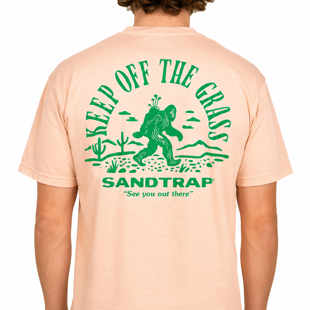 Male model wearing a peach Sandtrap T-shirt featuring a large green “Keep Off The Grass” graphic across the back. The design showcases a Bigfoot carrying golf clubs through a desert landscape with cacti, mountains, and the phrase “See you out there,” displayed against a clean studio background.