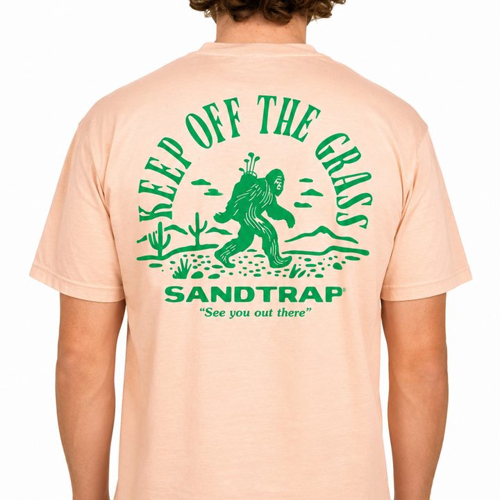 Male model wearing a peach Sandtrap T-shirt featuring a large green “Keep Off The Grass” graphic across the back. The design showcases a Bigfoot carrying golf clubs through a desert landscape with cacti, mountains, and the phrase “See you out there,” displayed against a clean studio background.