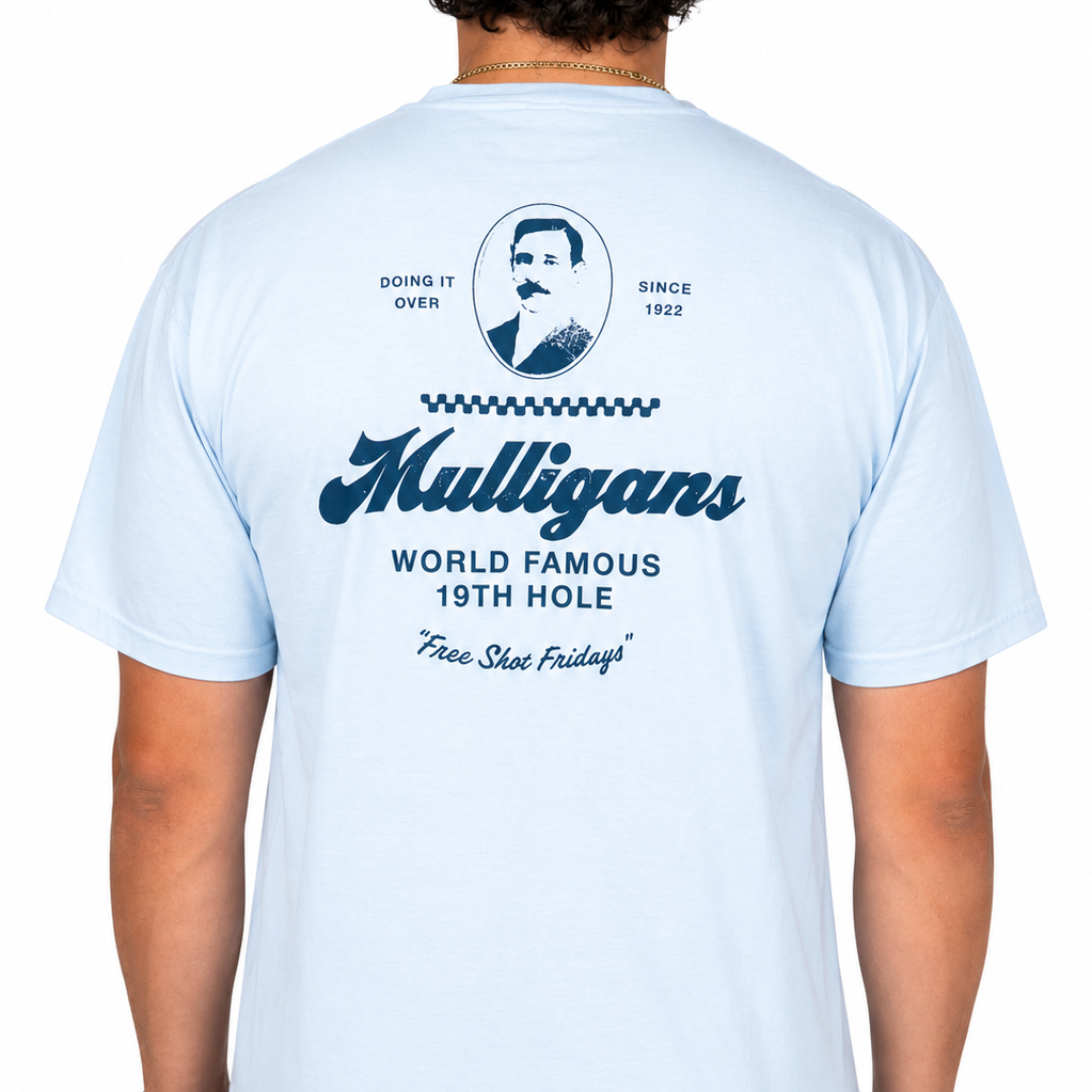 Male model wearing a light blue graphic t-shirt featuring a navy "Mulligans" design on the back with vintage portrait artwork, "World Famous 19th Hole" text, and "Free Shot Fridays" script against a clean white studio background.