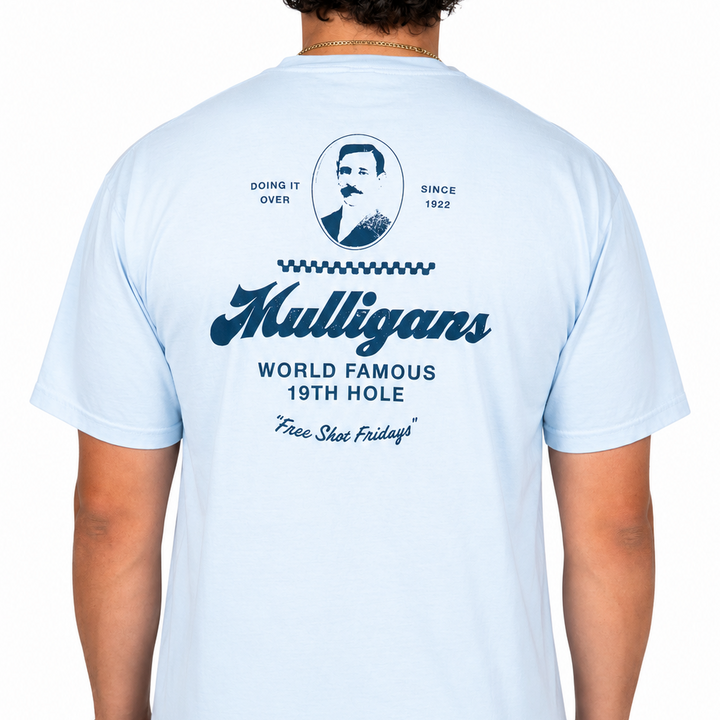 Male model wearing a light blue graphic t-shirt featuring a navy "Mulligans" design on the back with vintage portrait artwork, "World Famous 19th Hole" text, and "Free Shot Fridays" script against a clean white studio background.