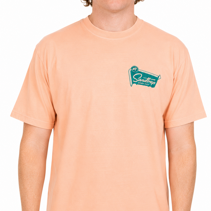 Male model wearing a peach Sandtrap t-shirt featuring a small teal Sandtrap Après Golf logo on the left chest against a clean white studio background.