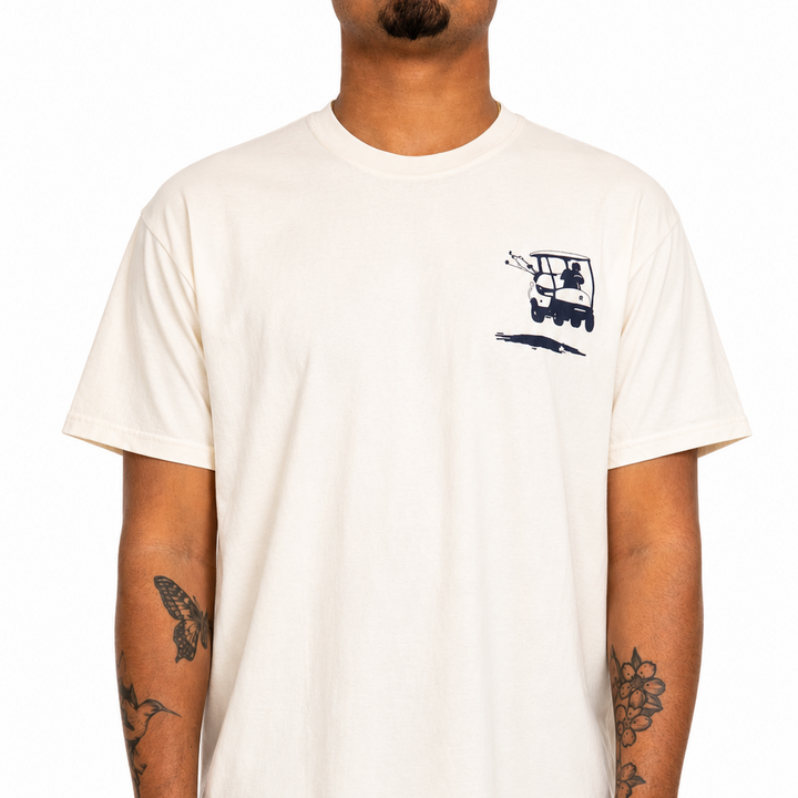 Male model wearing a cream Sandtrap Golf short-sleeve t-shirt featuring a navy golf cart graphic on the left chest. Relaxed fit crewneck tee shown against a clean white studio background.