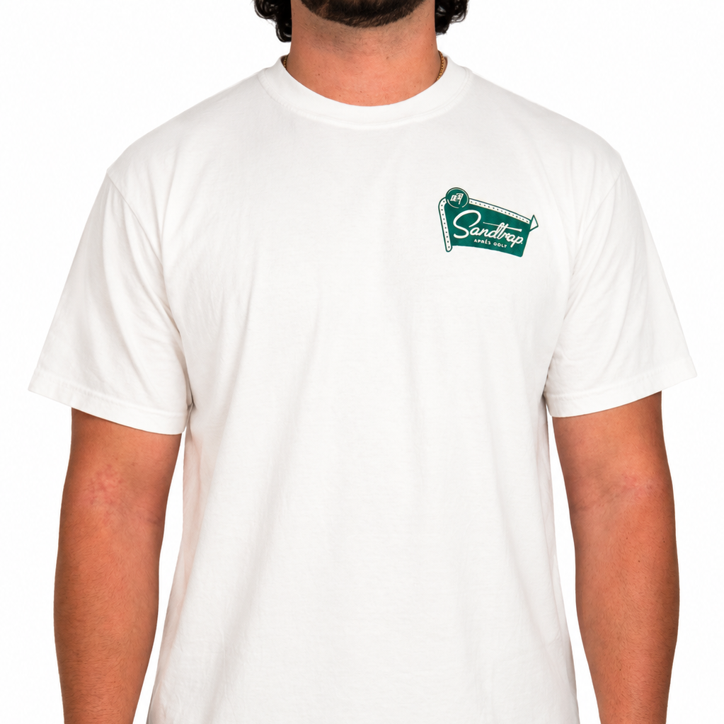 Male model wearing a white Sandtrap T-shirt featuring a small teal Sandtrap Hotel logo graphic on the left chest, paired with green shorts. Front-facing studio product image on a clean white background.