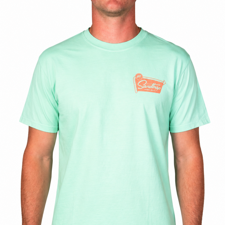 Male model wearing a mint green Sandtrap Après Golf t-shirt with a small coral logo graphic on the left chest product image on a clean white background.