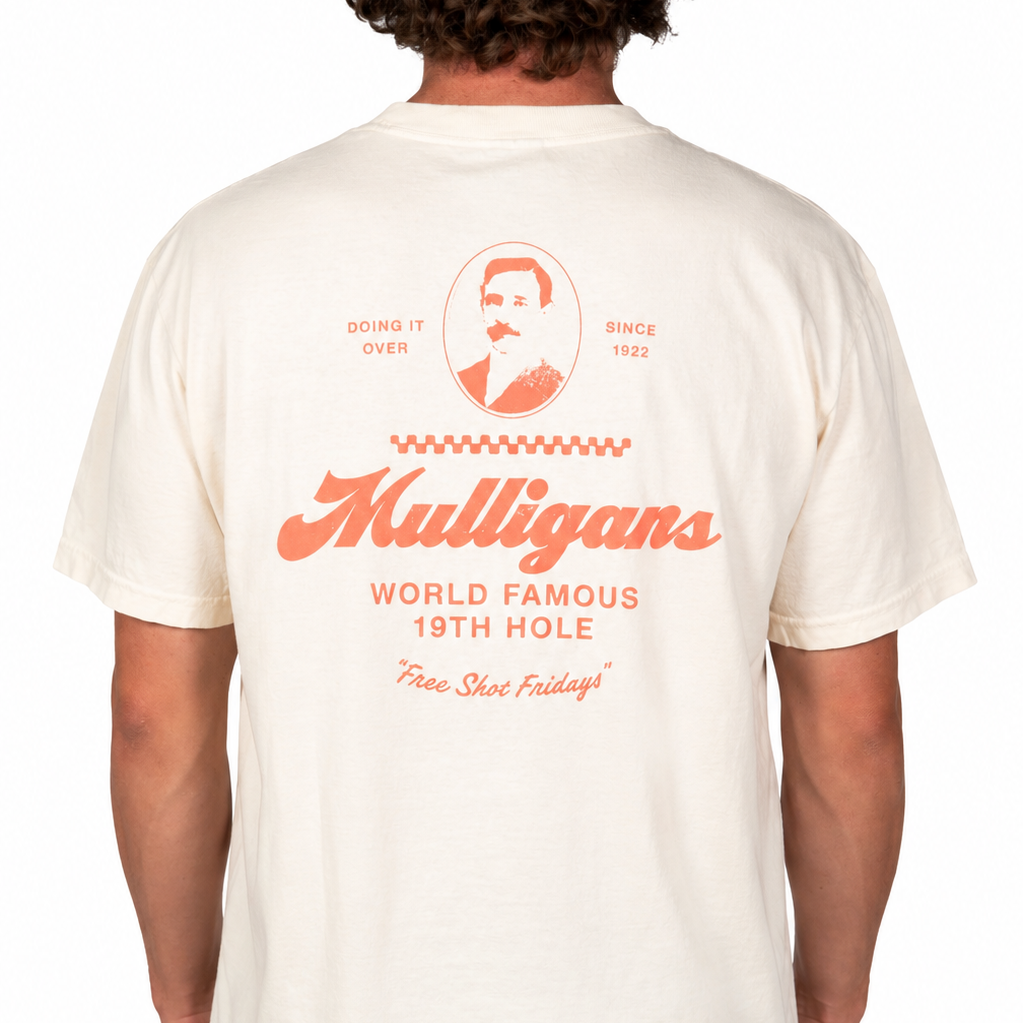 Male model wearing a cream-colored graphic t-shirt featuring a coral "Mulligans" design on the back with vintage-inspired typography, portrait artwork, and "World Famous 19th Hole" text against a clean white studio background.