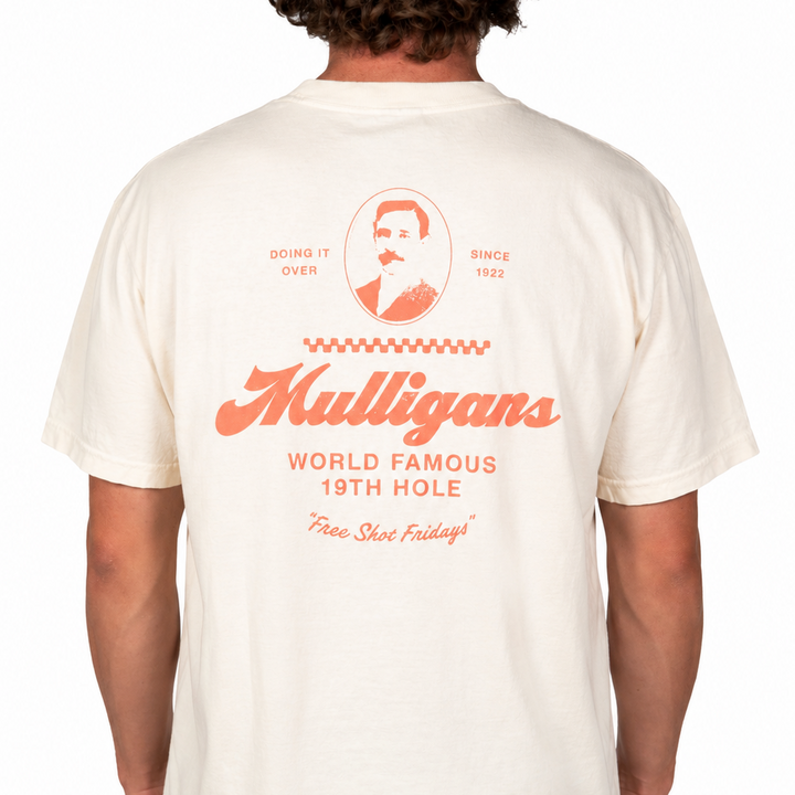 Male model wearing a cream-colored graphic t-shirt featuring a coral "Mulligans" design on the back with vintage-inspired typography, portrait artwork, and "World Famous 19th Hole" text against a clean white studio background.