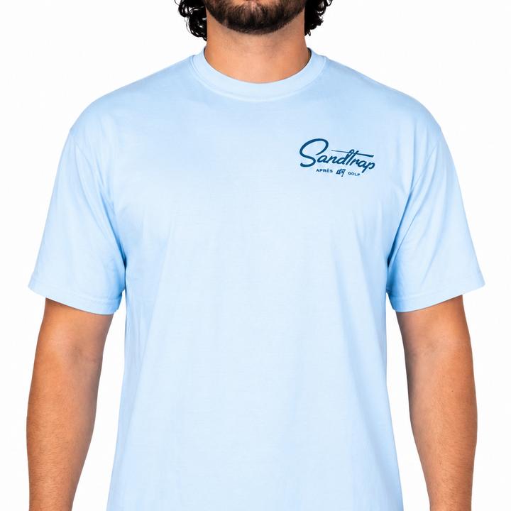 Male model wearing a light blue Sandtrap t-shirt featuring a small navy Sandtrap Après Golf logo on the left chest against a clean white studio background.