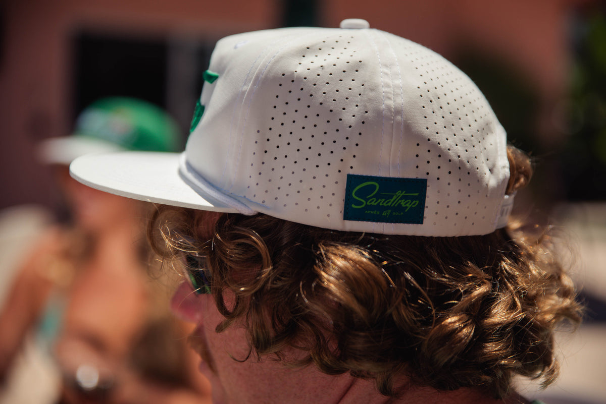 Person wearing a white cap with a Sandtrap logo, blurred background