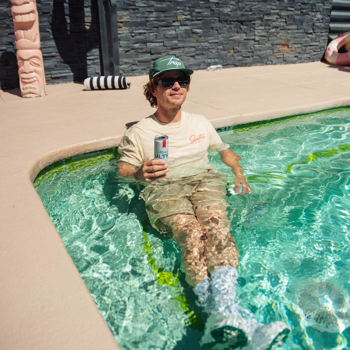 Male model relaxing in a hot tub with a desert cactus visible in the background, wearing a cream-colored Sandtrap t-shirt, creating a laid-back après-golf lifestyle scene.