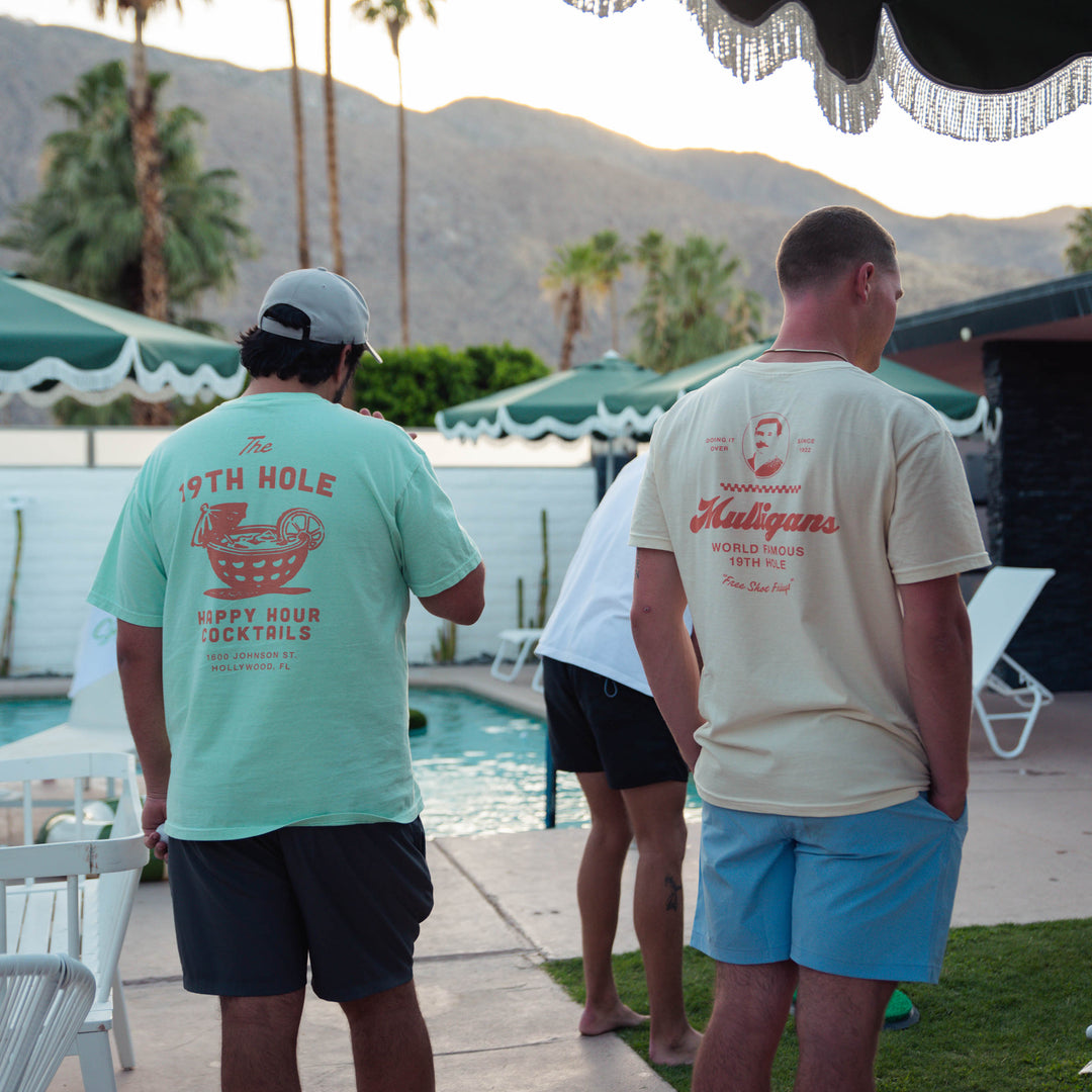 Two men standing by a poolside with palm trees and mountains in the background. Wearing The 19th Hole Happy Hour Cocktails” graphic on the back, with a cocktail-in-a-golf-ball illustration and Hollywood, Florida text.