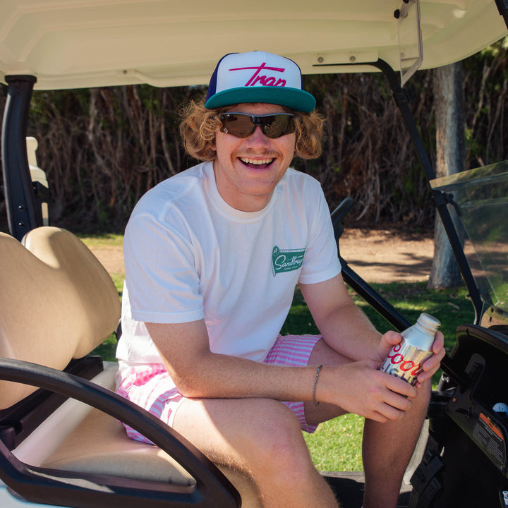 Male model sitting in a golf cart holding a drink, wearing sunglasses, Trap hat, and white Sandtrap T-shirt featuring a small teal Sandtrap Hotel logo graphic on the left chest.