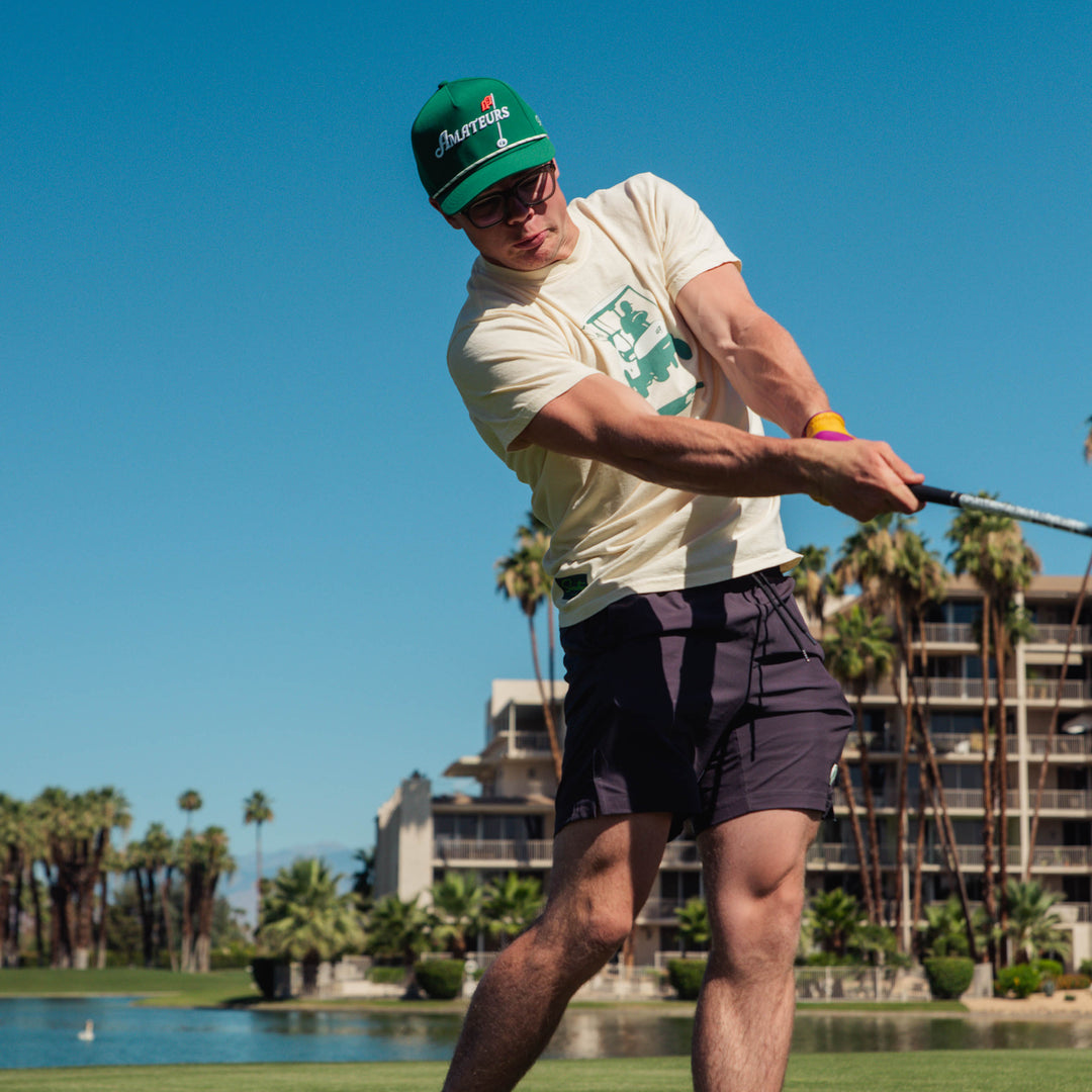Male model playing golf on a course with palm trees and buildings in the background wearing an off-white Sandtrap T-shirt featuring a teal golf cart graphic centered on the chest.
