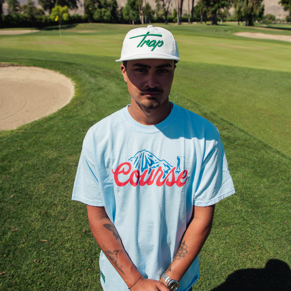 Male model wearing a Trap hat and t-shirt with a large red "Course" script graphic and blue mountain-and-golfer illustration centered on the chest.