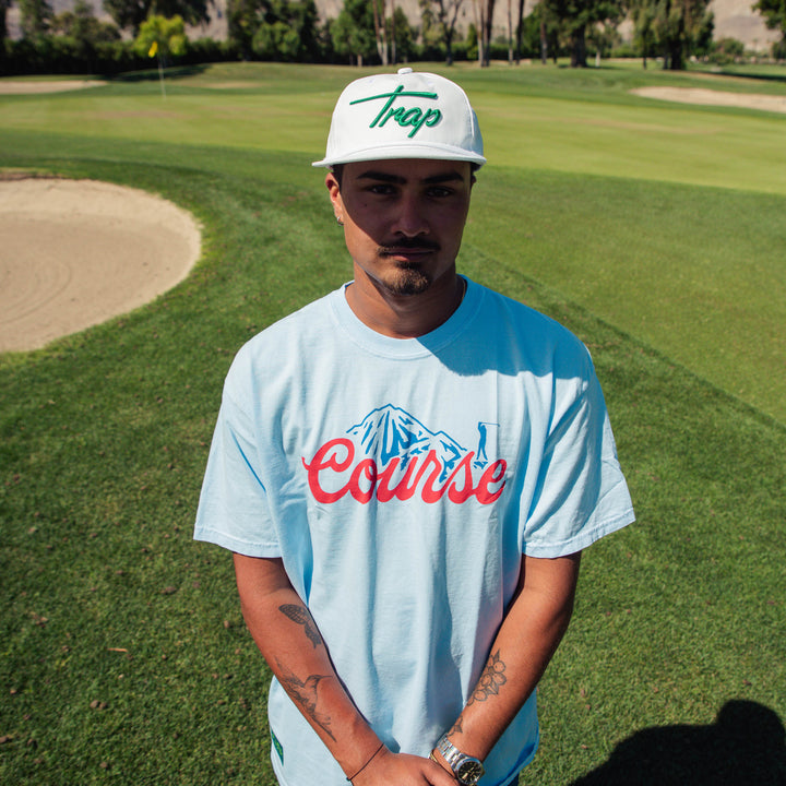 Male model wearing a Trap hat and t-shirt with a large red "Course" script graphic and blue mountain-and-golfer illustration centered on the chest.
