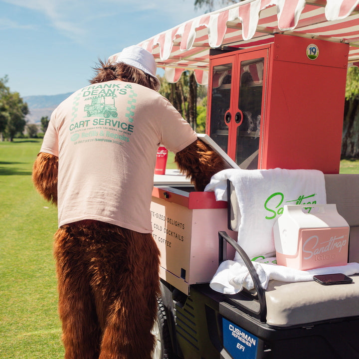 Person in a Sasquatch costume reaching into a golf cart bar setup on the course while wearing a peach Sandtrap T-shirt featuring the large “Frank &amp; Dean’s Cart Service” graphic on the back. The retro-inspired design showcases a vintage golf cart illustration, teal and mint green lettering, and playful repair shop-style details.
