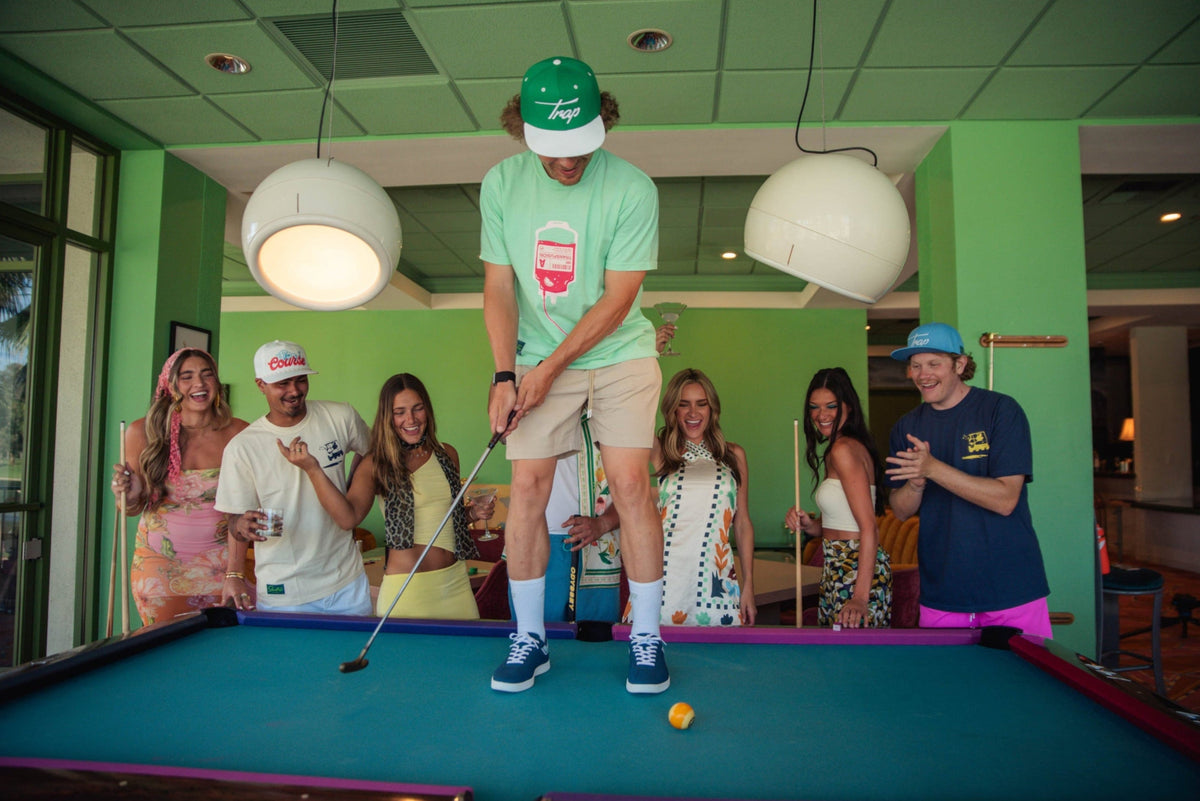 Group of people standing around a pool table in dressy attire watching guy on top of pool table putting with a pool ball. 