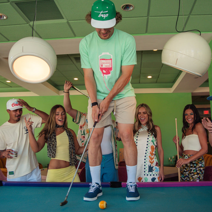 Male model standing on a pool table using a putter to hit a pool ball while a male and female models cheer him on. He is wearing a mint green graphic t-shirt featuring a pink and white transfusion IV bag design with tubing extending across the front.