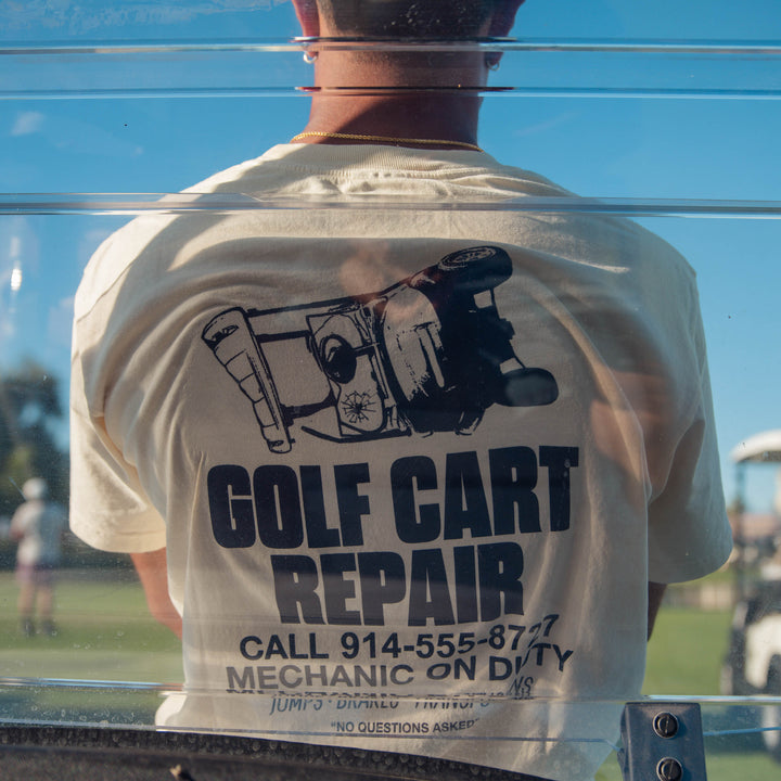 Male model wearing a cream Sandtrap Golf "Golf Cart Repair" graphic t-shirt with a large back print, leaning against the front of a golf cart on a golf course, showcasing the shirt.