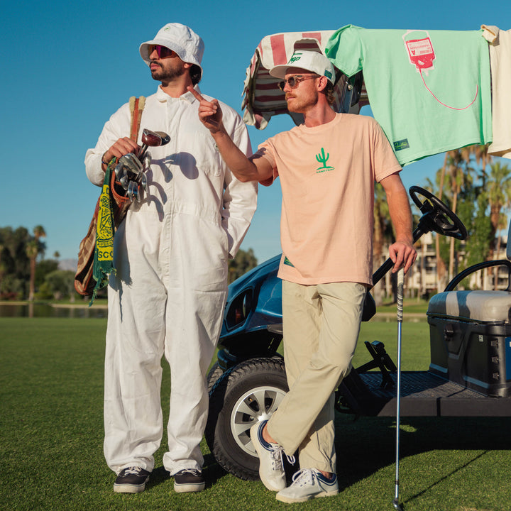 Two men on a golf course with one holding a golf club and the other standing next to a golf cart.