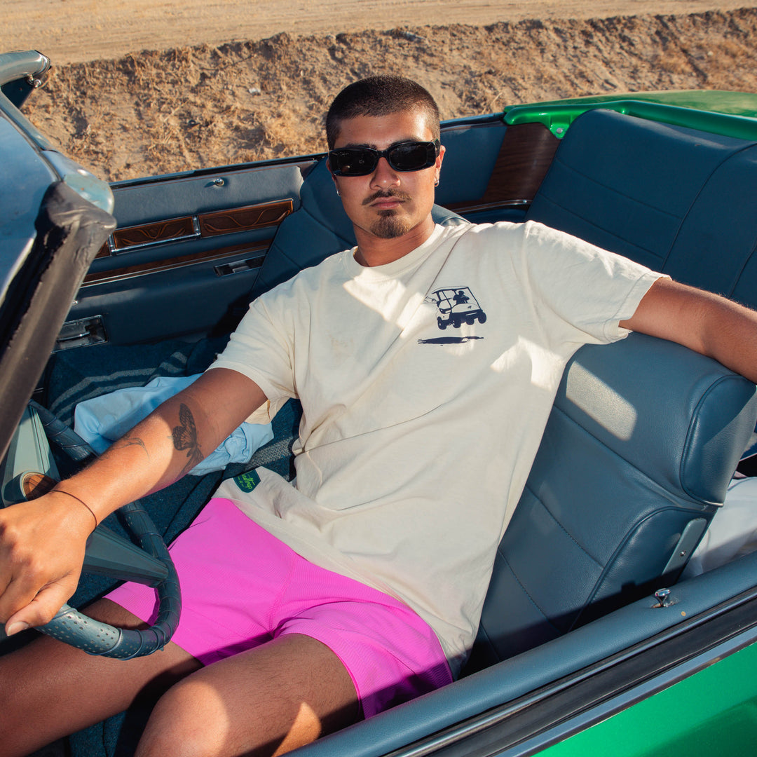 male model sitting in a convertible car in a desert setting wearing cream Sandtrap Golf short-sleeve t-shirt featuring a navy golf cart graphic on the left chest.