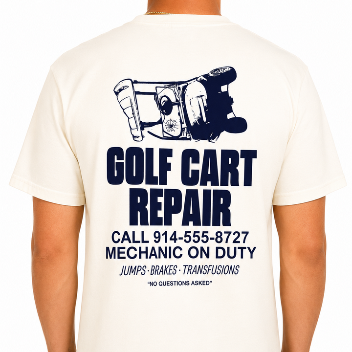 Male model wearing a cream Sandtrap Golf short-sleeve t-shirt featuring a large navy "Golf Cart Repair" graphic on the back with an overturned golf cart illustration and vintage repair shop-inspired text. Relaxed fit shown against a clean white studio background.