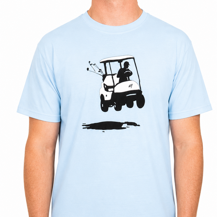 Male model wearing a light blue Sandtrap T-shirt featuring a centered golf cart graphic on the front. The minimalist design shows a golf cart with clubs in the back and a shadow beneath, displayed against a clean studio background.