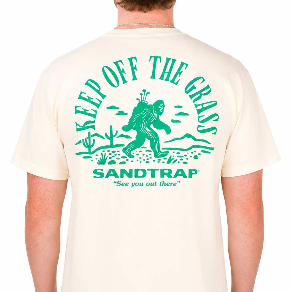 Male model wearing a cream Sandtrap T-shirt featuring a large green “Keep Off The Grass” graphic across the back. The design showcases a Bigfoot carrying golf clubs through a desert landscape with cacti, mountains, and Sandtrap branding, displayed against a clean studio background.