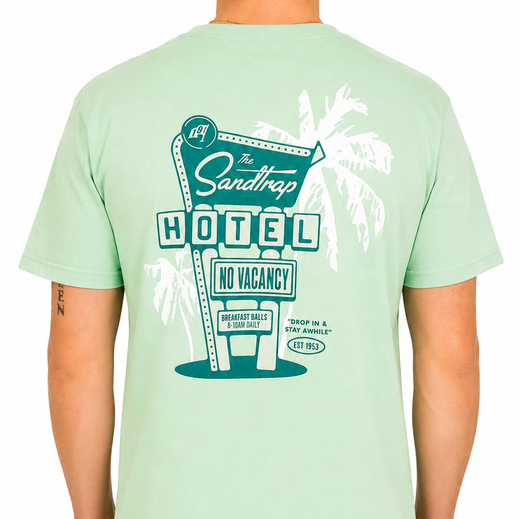 Male model shown from the back wearing a mint green Sandtrap Hotel graphic t-shirt. The large back print features a retro motel sign design with palm trees, “The Sandtrap Hotel,” “No Vacancy,” and golf-inspired details in teal and white, displayed against a clean white studio background.