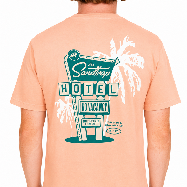 Back view of a peach Sandtrap t-shirt worn by a male model, featuring a large retro motel-inspired graphic with palm trees, a teal “Sandtrap Hotel” sign, “No Vacancy” lettering, and vintage resort-style details. Shown against a clean white studio background.