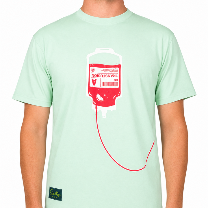 Front view of a male model wearing a mint green graphic t-shirt against a clean white studio background. The t-shirt features a large transfusion IV bag graphic in pink and white centered on the chest, with tubing extending across the lower front of the shirt.